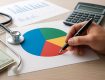 A hand holding a pen, drawing on a colorful pie chart with a stethoscope and calculator nearby, and a few dollar bills v