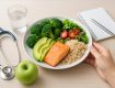 A healthy meal with salmon, brown rice, avocado, and vegetables on a plate, accompanied by an apple and a stethoscope