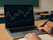 Close-up of a hand writing notes while looking at Forex trading charts on a laptop in a classroom setting